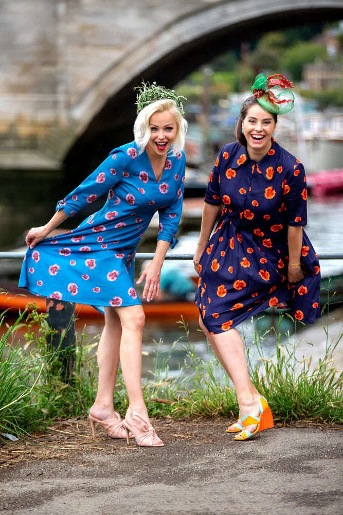 Two women wearing colourful floral dresses with fascinators by the riverside, vibrant summer fashion look