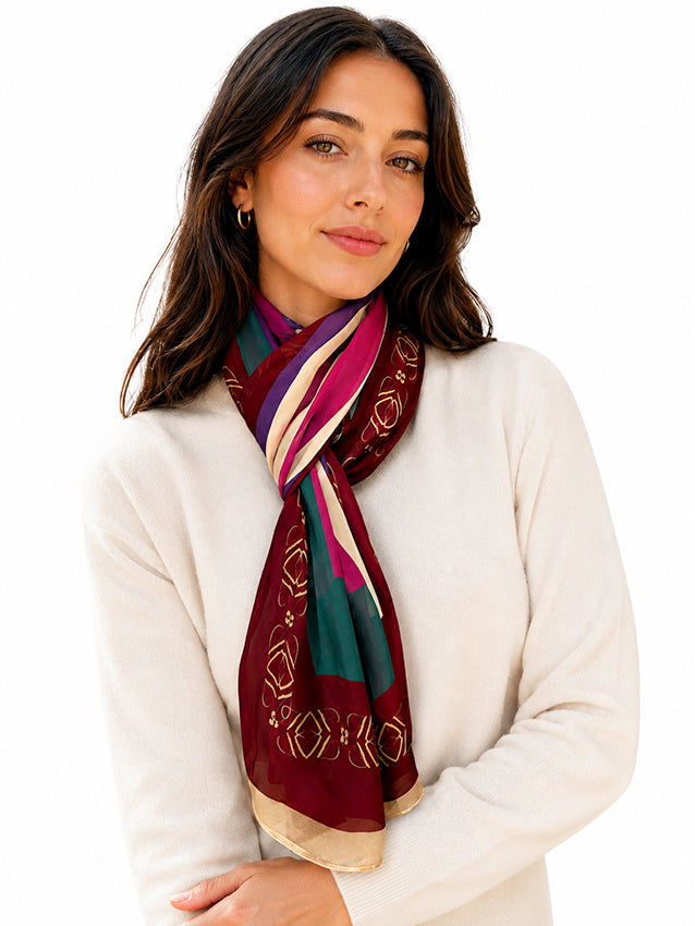 Elegant brunette model wearing a burgundy silk chiffon scarf with colourful geometric detailing and gold accents, styled with a soft ivory knit on a white background for timeless luxury womenswear by Moda de la Maria.
