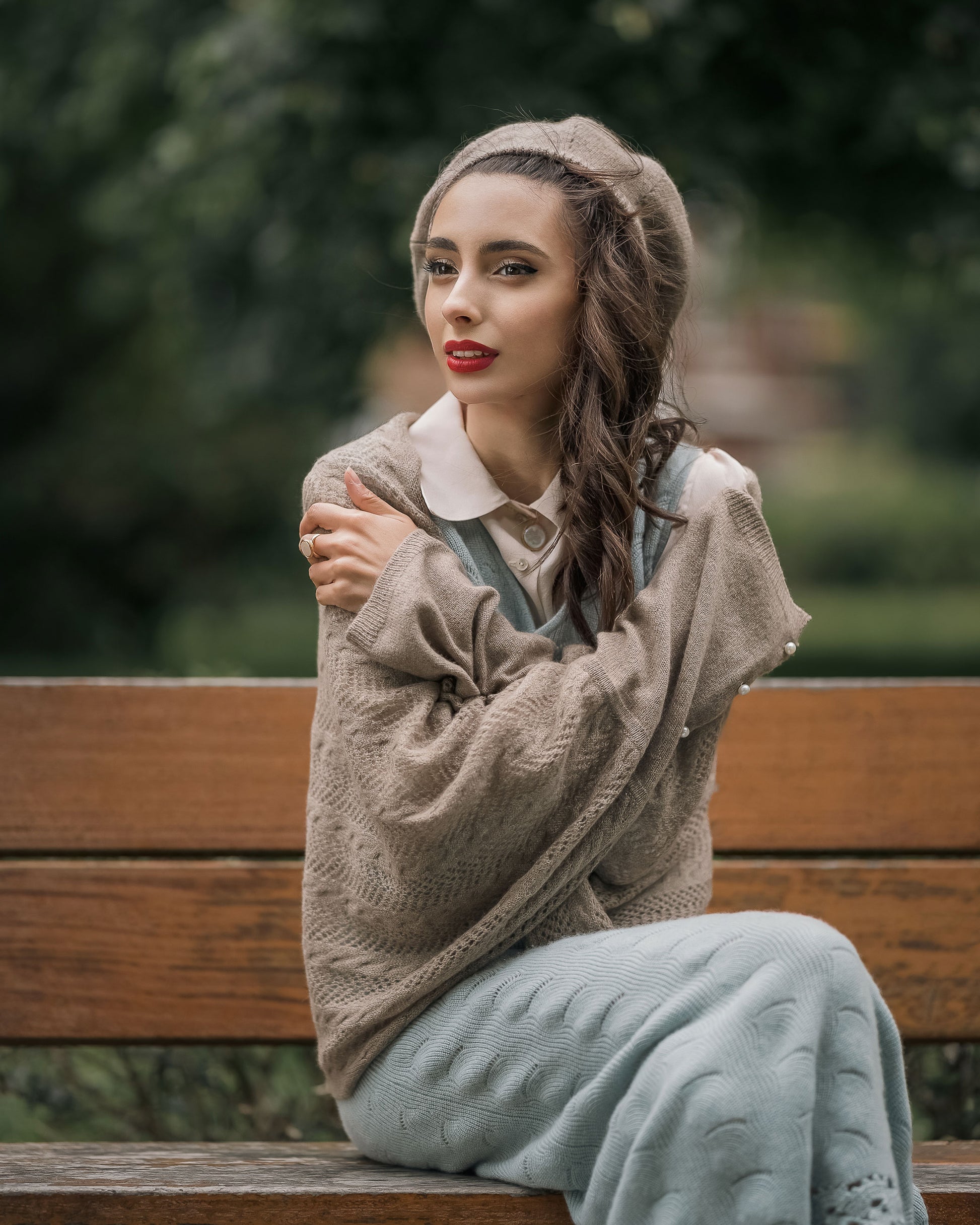 Side view of model on a park bench in a blue cashmere dress layered over a silk shirt, finished with scarf and hat, showcasing versatile styling by Moda de la Maria.